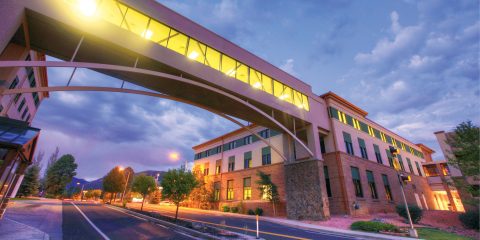 A building with an illuminated sky bridge, taken at dusk with a scenic sky and street below.