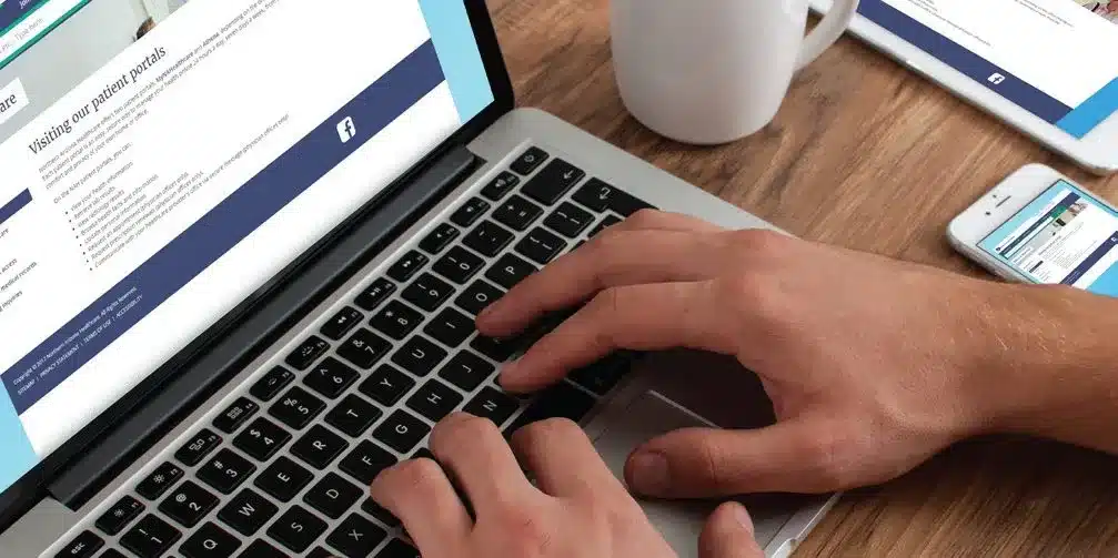 A person typing on a laptop, with a healthcare-related website on the screen. A smartphone and a cup of coffee sit on the wooden table beside the laptop.