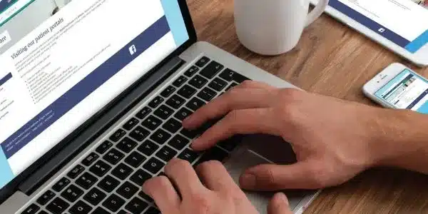 A person typing on a laptop, with a healthcare-related website on the screen. A smartphone and a cup of coffee sit on the wooden table beside the laptop.
