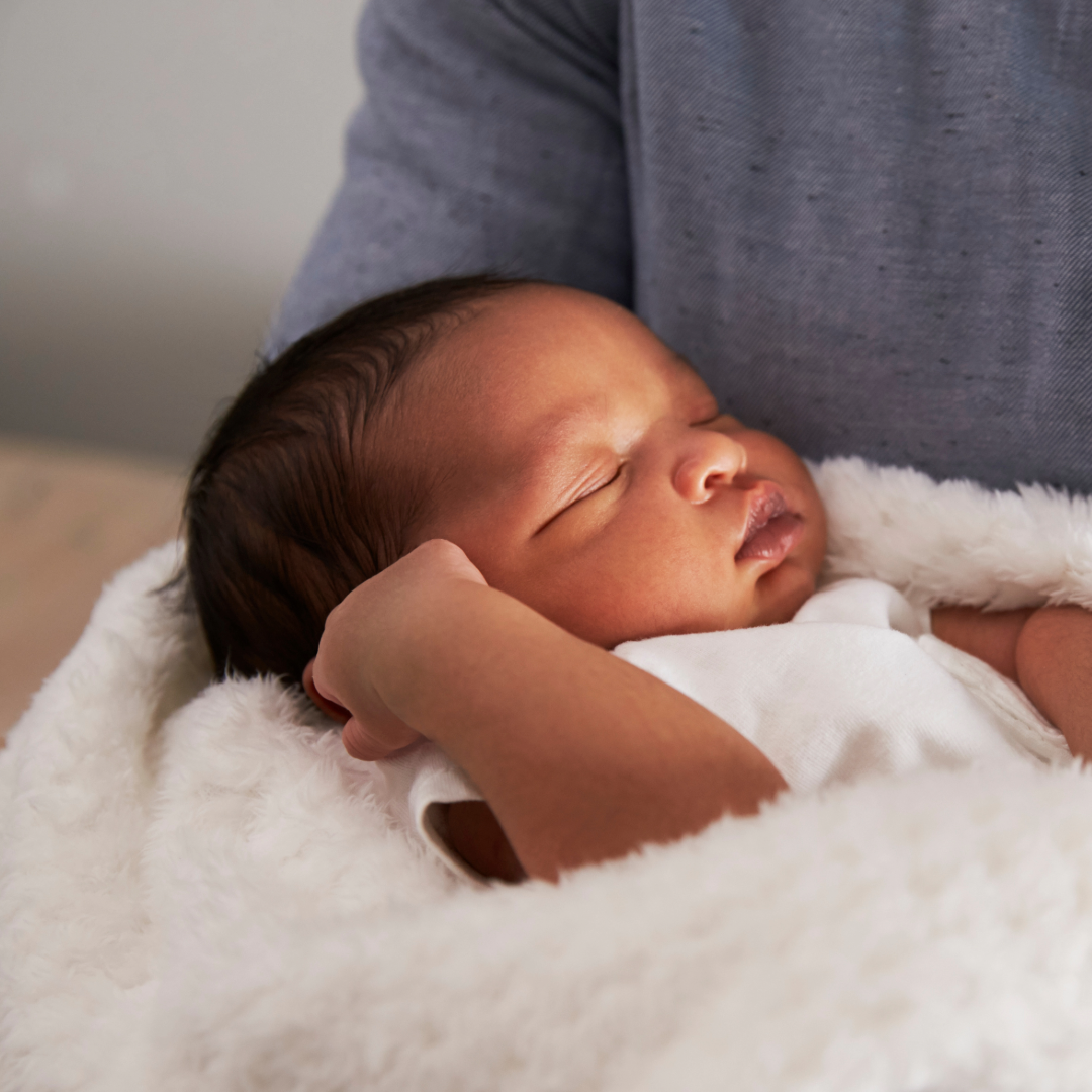 Sleeping newborn cradled in an adult’s arms, wrapped in a white blanket.