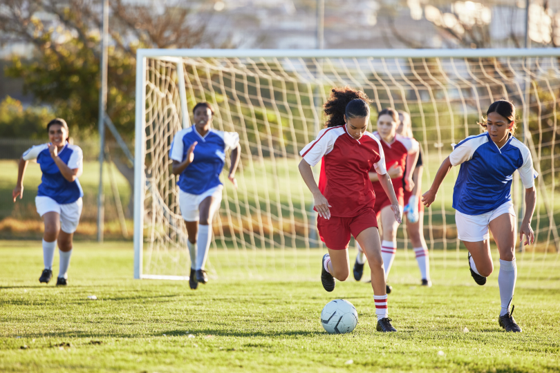 Girls playing soccer, with a player in red dribbling the ball as opponents in blue chase.