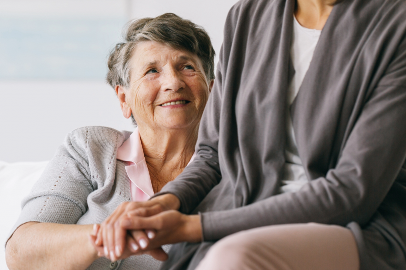 Older woman smiling and holding hands with another person, looking up at them indoors.