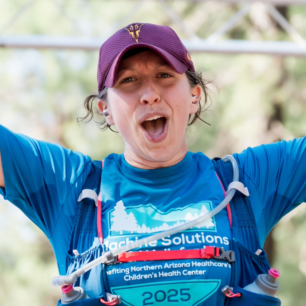 Person wearing a race bib and hydration vest, raising both arms in celebration at a running event.